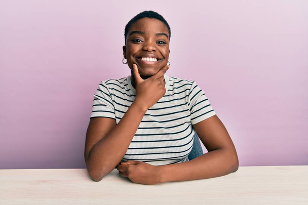 Νεαρή Αφροαμερικανή γυναίκα που φοράει καθημερινά ρούχα κάθεται στο τραπέζι και κοιτάζει με αυτοπεποίθηση την κάμερα χαμογελώντας με σταυρωμένα χέρια και το χέρι υψωμένο στο πηγούνι. θετική σκέψη.  - Φωτογραφία, εικόνα