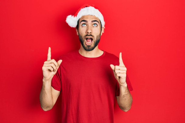 Jonge Spaanse man draagt kerstmuts verbaasd en verrast omhoog kijkend en wijzend met vingers en opgeheven armen.  - Foto, afbeelding