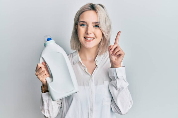 Młoda blondynka trzymająca butelkę detergentu zaskoczona pomysłem lub pytaniem wskazującym palcem szczęśliwą twarz, numer jeden  - Zdjęcie, obraz