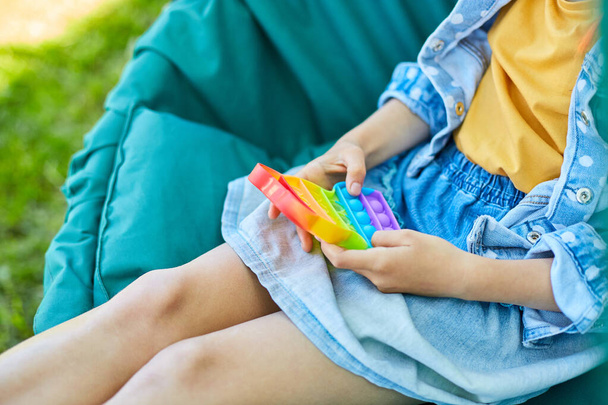 A little girl the on hanging chair outdoors play pop it, kid hands playing playing bubbles of a rainbow destressing toy, fidget toy in backyard of house on a sunny summer day, summer time vacation. - Фото, изображение