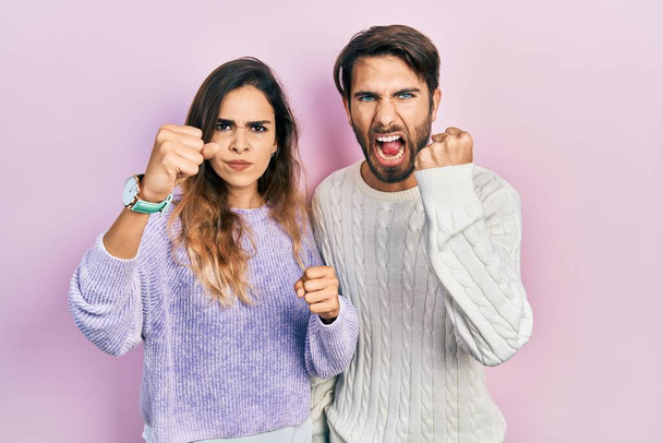 Νεαρό ζευγάρι ισπανόφωνων που φορούν καθημερινά ρούχα θυμωμένοι και θυμωμένοι υψώνοντας γροθιές απογοητευμένοι και εξοργισμένοι ενώ φωνάζουν με θυμό. οργή και επιθετική αντίληψη.  - Φωτογραφία, εικόνα