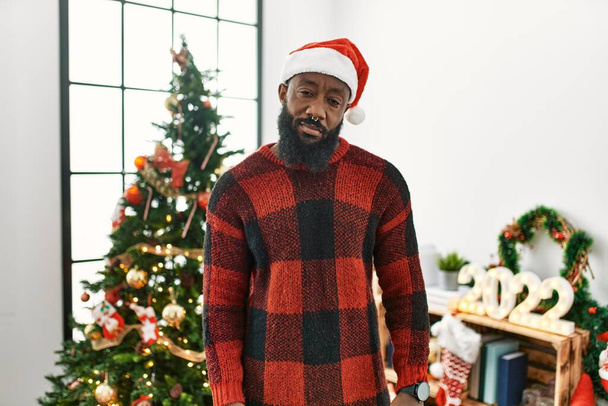 Αφροαμερικάνος που φοράει καπέλο Σάντα Κλάους στέκεται δίπλα στο χριστουγεννιάτικο δέντρο δείχνοντας νυσταγμένος και κουρασμένος, εξαντλημένος από την κούραση και το μεθύσι, τεμπέλης μάτια το πρωί.  - Φωτογραφία, εικόνα