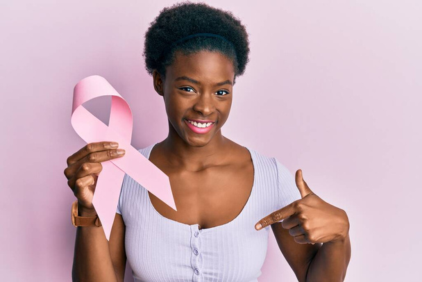 Young african american girl holding pink cancer ribbon pointing finger to one self smiling happy and proud  - Foto, Bild