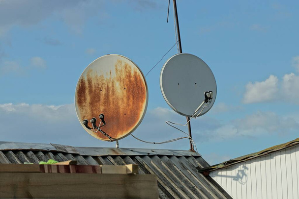 дві сірі круглі супутникові тарілки в коричневій іржі на даху шиферу приватного будинку на тлі блакитного неба
 - Фото, зображення