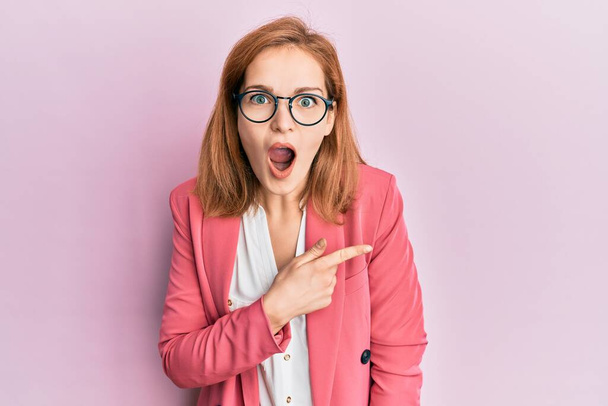 Young caucasian woman wearing business style and glasses surprised pointing with finger to the side, open mouth amazed expression.  - Foto, Bild