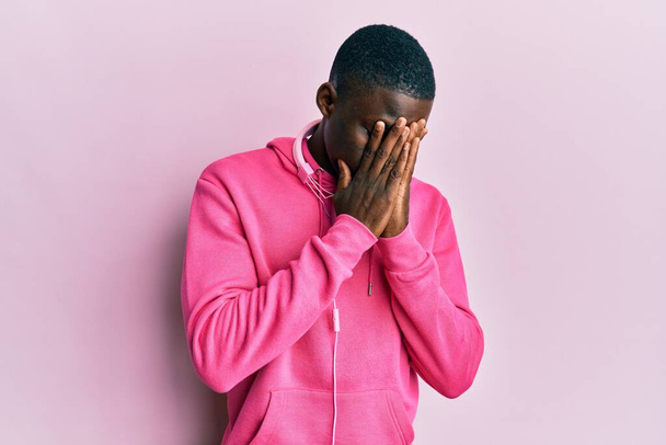 Jonge Afro-Amerikaanse man draagt sportkleding en gebruikt koptelefoons met een droevige uitdrukking die gezicht met handen bedekken terwijl hij huilt. depressie concept.  - Foto, afbeelding