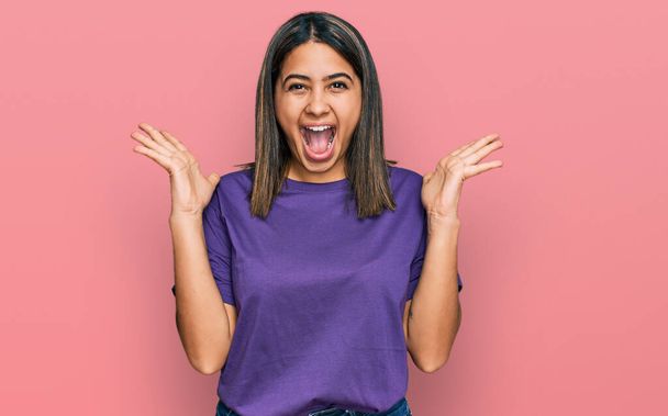 Jeune fille hispanique portant t-shirt violet occasionnel célébrant fou et étonné pour le succès avec les bras levés et les yeux ouverts hurlant excité. concept gagnant  - Photo, image