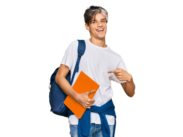 Молодий іспаномовний чоловік носить студентський рюкзак і тримає книги усміхнені щасливі вказуючи рукою і пальцем
  - Фото, зображення