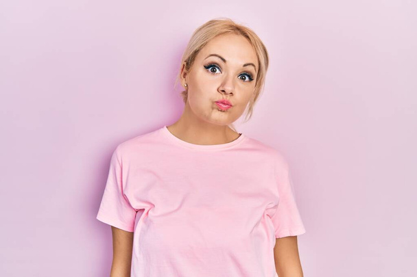 Jeune femme blonde portant un t-shirt rose décontracté gonflant les joues avec un visage drôle. bouche gonflée d'air, expression folle.  - Photo, image
