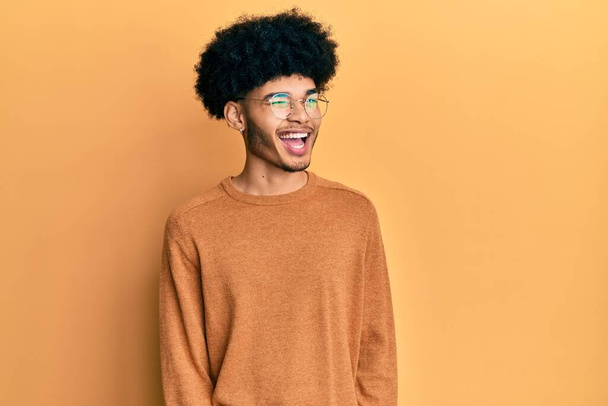 Junger afrikanisch-amerikanischer Mann mit Afro-Haaren in lässigem Winterpullover zwinkernd in die Kamera blickend mit sexy Ausdruck, fröhlichem und glücklichem Gesicht.  - Foto, Bild