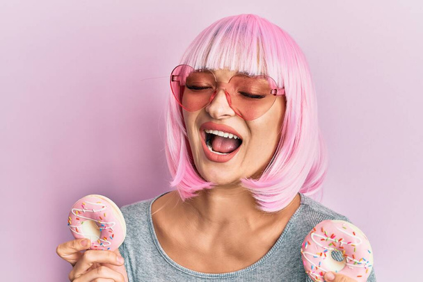 Junge kaukasische Frau mit pinkfarbener Perücke hält Donuts lächelnd in der Hand und lacht laut, weil lustig verrückter Witz.  - Foto, Bild