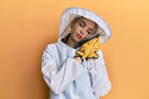 Όμορφη ξανθιά καυκάσια γυναίκα φορώντας προστατευτική στολή μελισσοκόμου κοιμάται κουρασμένος ονειρεύεται και ποζάρουν με τα χέρια μαζί, ενώ χαμογελά με κλειστά μάτια.  - Φωτογραφία, εικόνα