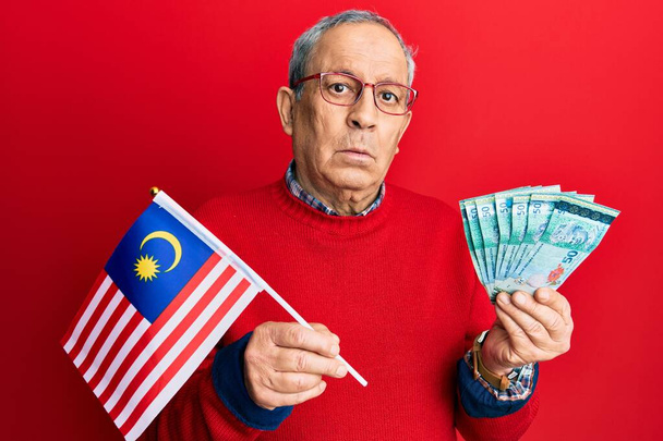 Knappe oudere man met grijs haar die de vlag van Maleisië vasthield en Maleisische ringgit bankbiljetten zonder enig idee en verwarde uitdrukking. twijfelconcept.  - Foto, afbeelding