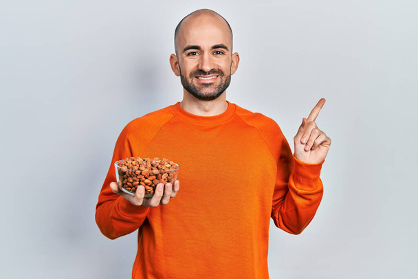 Young bald man holding peanuts smiling happy pointing with hand and finger to the side  - Foto, Imagem
