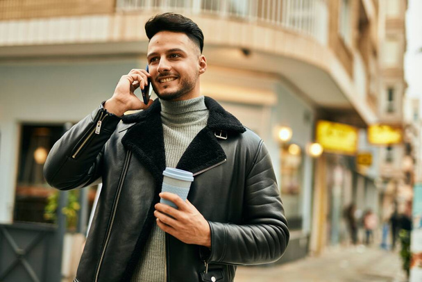 Νεαρός Άραβας μιλάει στο smartphone πίνοντας καφέ στην πόλη. - Φωτογραφία, εικόνα