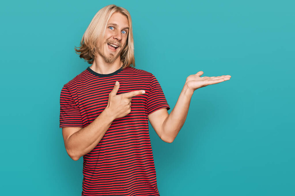 Белый мужчина с длинными светлыми волосами в повседневной полосатой футболке изумлен и улыбается в камеру, показывая рукой и указывая пальцем.  - Фото, изображение