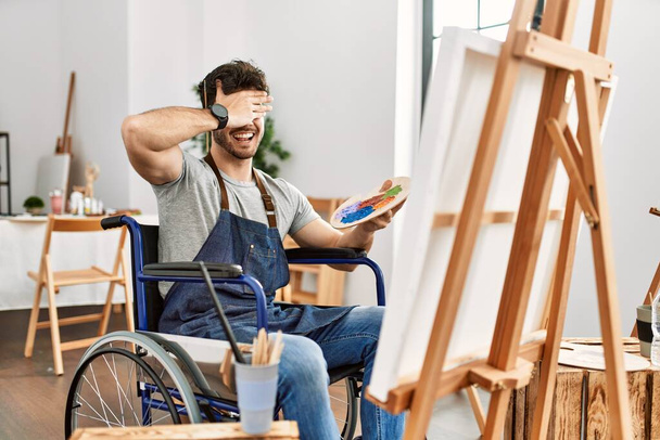 Junger hispanischer Mann, der im Kunstatelier im Rollstuhl sitzt, lächelt und lacht mit der Hand auf dem Gesicht und verdeckt die Augen zur Überraschung. Blindes Konzept.  - Foto, Bild