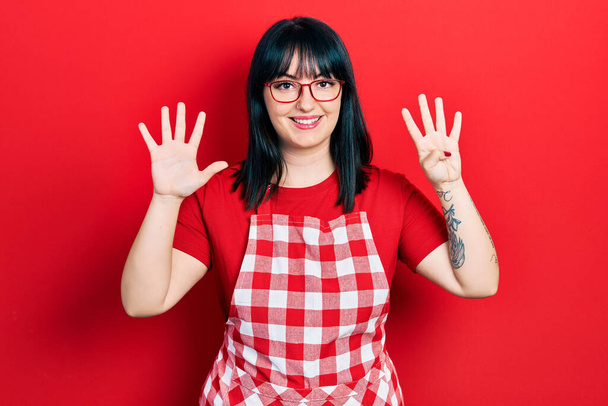Giovane donna ispanica indossa grembiule cuoco e occhiali mostrando e indicando con le dita numero nove, sorridendo fiducioso e felice.  - Foto, immagini