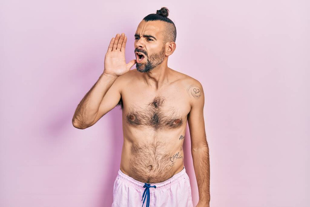 Jonge Spaanse man zonder shirt in zwempak schreeuwend en schreeuwend van hand op mond. communicatieconcept.  - Foto, afbeelding