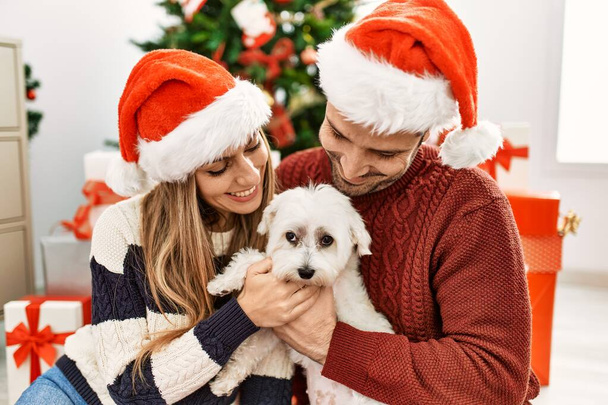 Młoda latynoska para w świątecznym kapeluszu siedząca na podłodze z psem w domu. - Zdjęcie, obraz