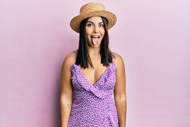 Young brunette woman wearing summer dress and hat sticking tongue out happy with funny expression. emotion concept.  - Photo, Image