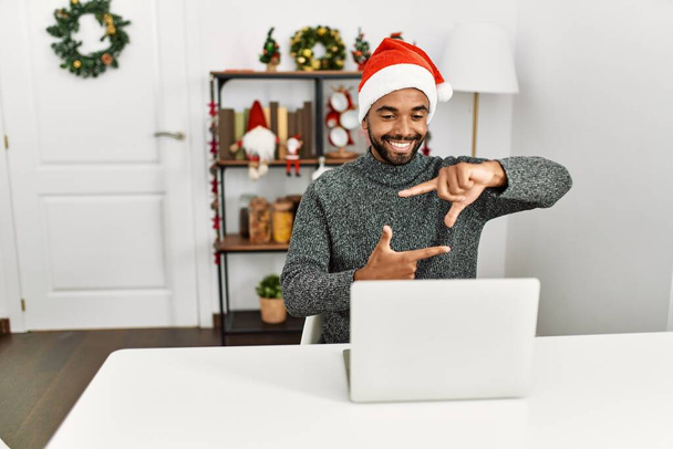 Молодий іспаномовний чоловік з бородою в різдвяному капелюсі використовує ноутбук, посміхаючись роблячи рамку руками і пальцями з щасливим обличчям. креативність і концепція фотографії
.  - Фото, зображення