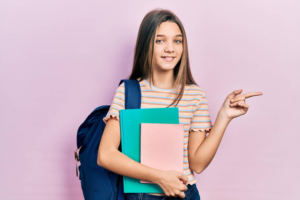 Jeune fille brune tenant sac à dos étudiant et livres souriant heureux pointant avec la main et le doigt sur le côté  - Photo, image
