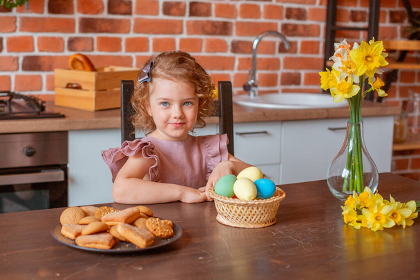 маленькая милая девочка с корзиной пасхальных яиц на кухне дома - Фото, изображение