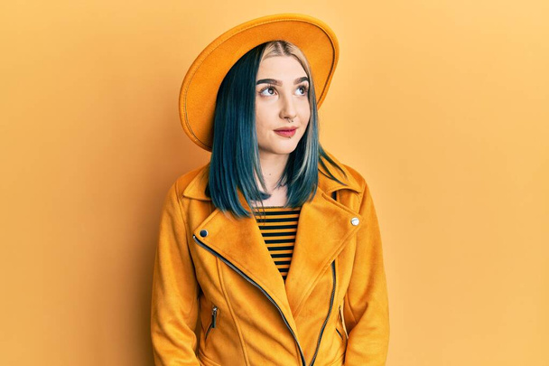 Nuori moderni tyttö yllään keltainen hattu ja nahkatakki hymyillen sivuun ja tuijottaen pois ajattelu.  - Valokuva, kuva