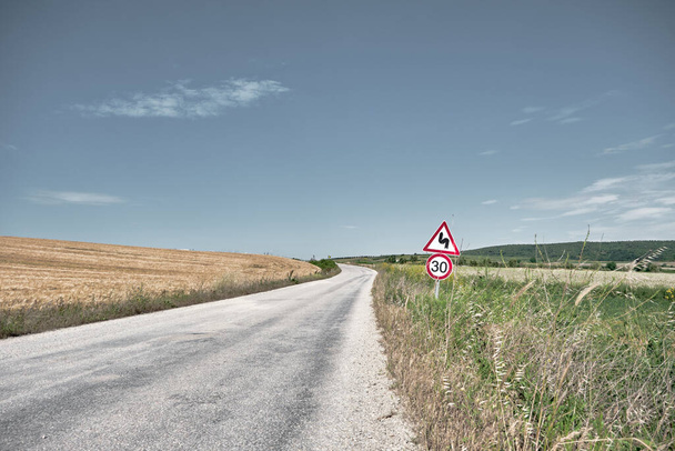Δρόμος και υπέροχη κίτρινο σιτάρι μούρα γεωργικές εκτάσεις κοντά στο δρόμο με θολό και ανοιχτό φόντο του ουρανού. Οδική σήμανση δίπλα στην οδό για την καμπύλη S και 30 km / h περιορισμός ταχύτητας - Φωτογραφία, εικόνα
