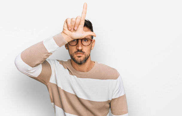 Handsome man with beard wearing casual clothes and glasses making fun of people with fingers on forehead doing loser gesture mocking and insulting.  - Photo, Image