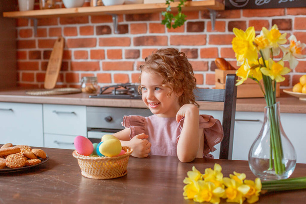 маленькая милая девочка с корзиной пасхальных яиц на кухне дома - Фото, изображение