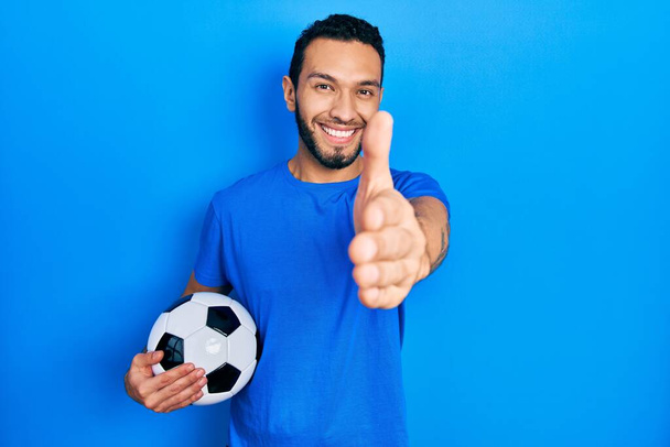 Latynos z brodą trzymający piłkę nożną uśmiechnięty, przyjazny, oferujący uścisk dłoni jako powitanie i powitanie. udany biznes.  - Zdjęcie, obraz