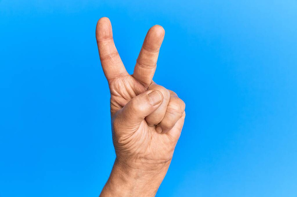 Hand eines hochrangigen hispanischen Mannes über blauem, isoliertem Hintergrund, der die Zahl 2 zählt, die zwei Finger zeigt, Sieg und Siegersymbol gestikuliert  - Foto, Bild