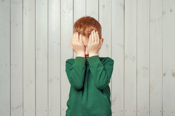 Δυστυχισμένο κοκκινομάλλικο αγοράκι 7-9 ετών κλαίει με τα χέρια του να καλύπτουν το πρόσωπό του, στέκεται πάνω σε ξύλινο φόντο - Φωτογραφία, εικόνα