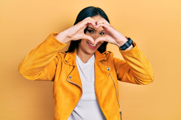 Красивая латиноамериканка с пирсингом носа в желтой кожаной куртке делает форму сердца с рукой и пальцами, улыбаясь, глядя сквозь знак  - Фото, изображение