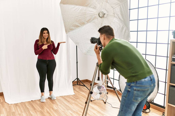 Młoda kobieta w studiu fotograficznym zdumiona i uśmiechnięta do aparatu prezentując się z ręką i wskazując palcem.  - Zdjęcie, obraz