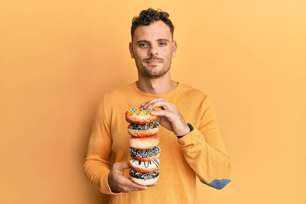 Νεαρός Ισπανόφωνος κρατώντας νόστιμα πολύχρωμα ντόνατς χαλαρά με σοβαρή έκφραση στο πρόσωπο. απλή και φυσική κοιτάζοντας την κάμερα.  - Φωτογραφία, εικόνα