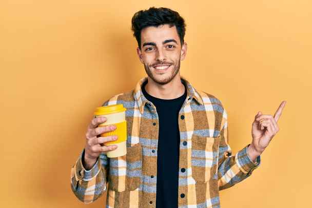 Junger hispanischer Mann hält Kaffee und lächelt glücklich und zeigt mit Hand und Finger zur Seite  - Foto, Bild