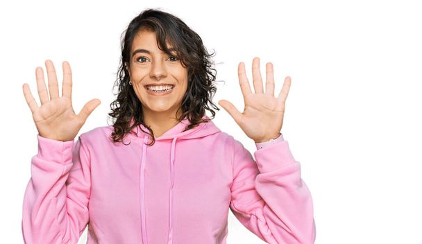 Νεαρή Ισπανίδα γυναίκα φορώντας casual φούτερ δείχνει και δείχνει προς τα πάνω με τα δάχτυλα νούμερο δέκα, ενώ χαμογελά αυτοπεποίθηση και χαρούμενος.  - Φωτογραφία, εικόνα