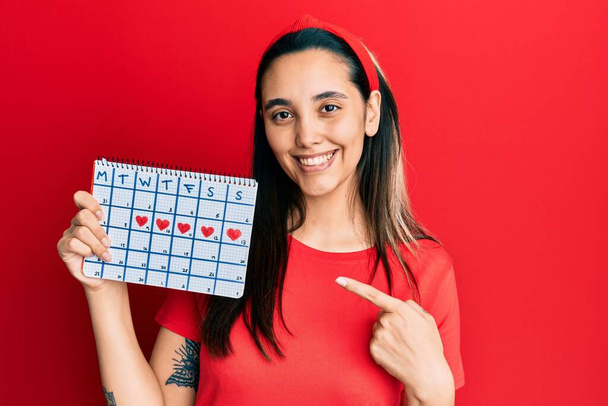 Joven mujer hispana sosteniendo el calendario del corazón sonriendo feliz señalando con la mano y el dedo  - Foto, Imagen