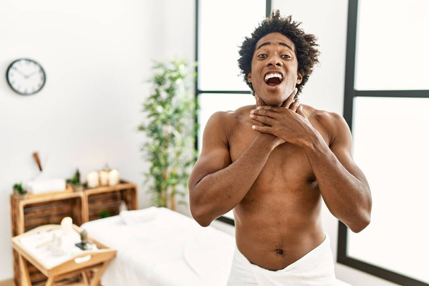 Jonge Afro-Amerikaanse man zonder shirt die een handdoek draagt in een schoonheidscentrum schreeuwend en verstikkend omdat hij gewurgd is. gezondheidsproblemen. verstikking en zelfmoordconcept.  - Foto, afbeelding