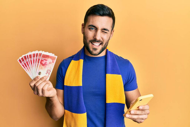 Młody Hiszpan kibic kibicujący piłce nożnej za pomocą smartfona trzymającego izraelskie szekle mrugające banknoty patrzące w kamerę z seksowną ekspresją, wesołą i szczęśliwą twarzą.  - Zdjęcie, obraz