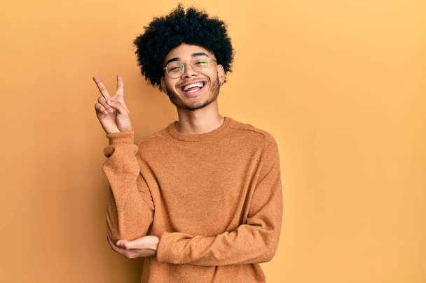 Junger afrikanisch-amerikanischer Mann mit Afro-Haaren in lässigem Winterpullover, lächelnd mit glücklichem Gesicht, das in die Kamera zwinkert und das Siegeszeichen macht. Nummer zwei.  - Foto, Bild