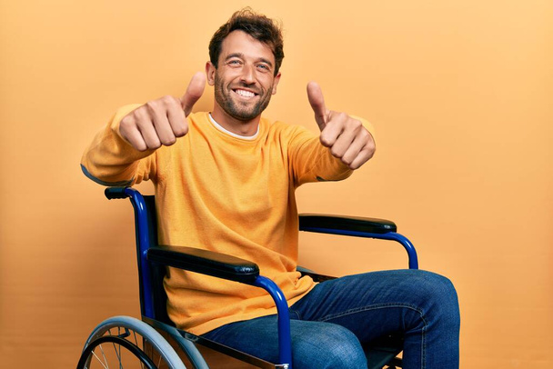 Schöner Mann mit Bart, der im Rollstuhl sitzt und eine positive Geste mit der Hand macht, Daumen hoch lächelnd und glücklich über den Erfolg. Siegergeste.  - Foto, Bild