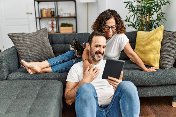 Middle age hispanic couple smiling happy and using touchpad. Sitting on the sofa with dogs at home. - Photo, Image