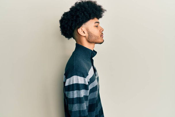 Jeune homme afro-américain avec des cheveux afro portant des vêtements décontractés regardant vers le côté, pose de profil relax avec visage naturel avec sourire confiant.  - Photo, image