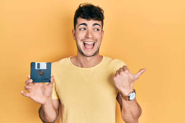 Giovane uomo ispanico in possesso di floppy disk che punta il pollice verso l'alto per il lato sorridente felice con la bocca aperta  - Foto, immagini