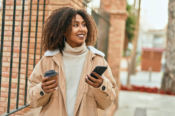 アフリカ系アメリカ人の若い女性が市内でスマートフォンやコーヒーを飲んでいる. - 写真・画像