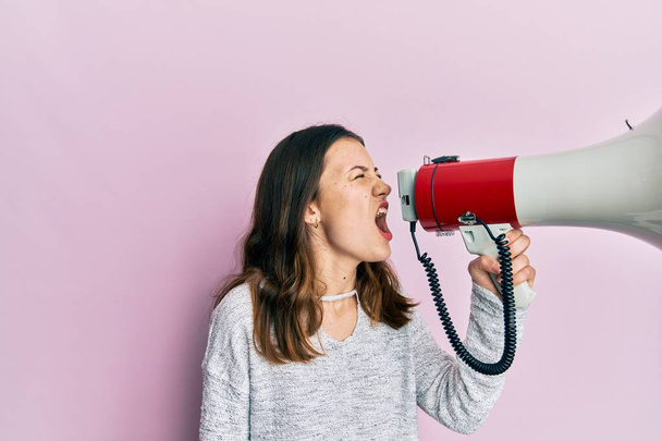Young brunette woman shouting and screaming through megaphone over pink isolated background - 写真・画像